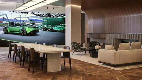 An Aston Martin lounge with a stone dining table, armchairs and a sofa under a curved walnut ceiling, beside a large screen showing a green DB12 next to the Aston Martin Formula One car.