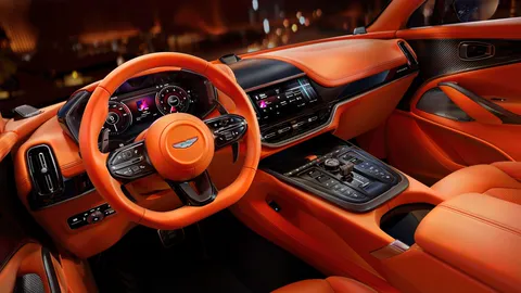Aston Martin DB12 interior in orange leather: the steering wheel, digital instrument cluster, central infotainment touchscreen and centre console switchgear.