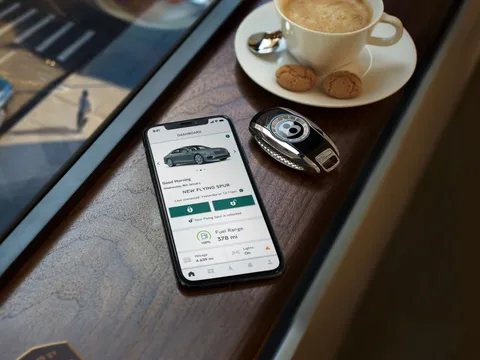 The My Bentley app on a phone beside a Bentley key and a cup of coffee on walnut veneer, its home screen greeting the owner.